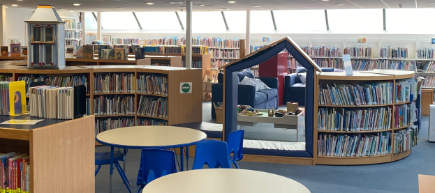 Eine Bibliothek mit Bücherregalen, Tischen, Stühlen, Sofas mit Kissen, einer Säule und Deckenleuchten.