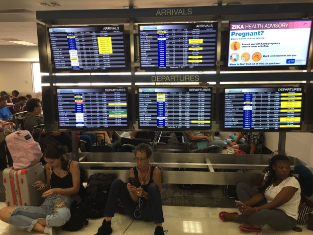Eine Gruppe von Menschen sitzt auf dem Boden eines Flughafens, einige halten Handys, mit verstreuten Taschen und Zika-Virus-Informationen im Hintergrund.