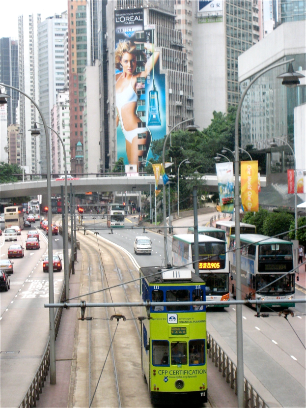 Eine belebte Straße in der Stadt mit verschiedenen Fahrzeugen, darunter Doppeldeckerbusse, Autos und andere Fahrzeuge, umgeben von hohen Gebäuden, Masten, Lichtern, Schildern, Bannern, Bäumen und einer Brücke unter einem klaren blauen Himmel, mit Text unten.
