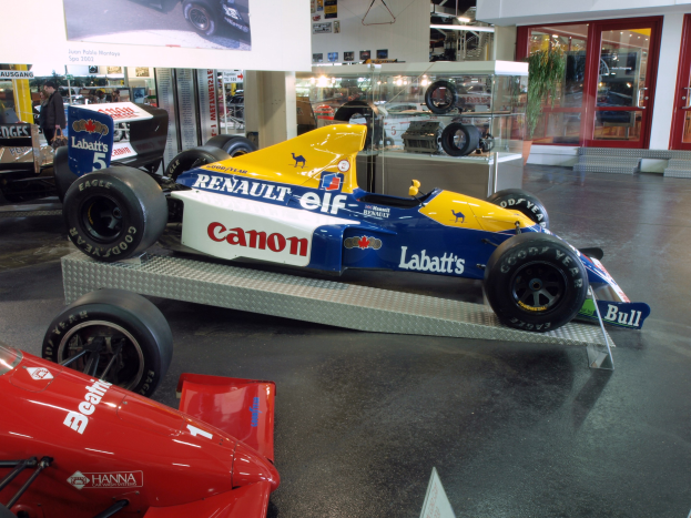 Eine Museumsausstellung zeigt verschiedene Rennwagen, darunter ein Renault-Formel-1-Auto, mit Besuchern, die die Ausstellungsstücke betrachten und eine Texttafel an der Wand im Hintergrund.