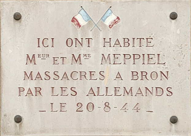 Steintafel an einer Gebäudefassade mit zwei Flaggen, beschriftet mit Text, der ein Massaker gedenkt.