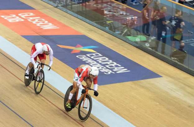 Zwei Männer mit Helmen fahren auf einer Indoor-Bahn vor einem Publikum, mit einer Glaswand, die 'Europameisterschaften Glasgow 2018' anzeigt und Zuschauern dahinter.