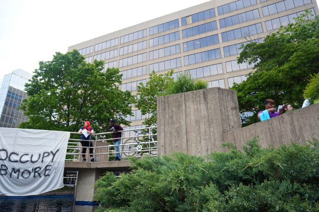 Menschen auf einer Brücke mit einem Banner und einem Fahrrad, mit Wasser darunter, Bäumen, Gebäuden und Himmel im Hintergrund.