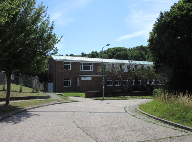 Ein Schulgebäude mit Fenstern, einem Straßenmast, einem Schild, einer Gruppe von Bäumen, Gras, einem Zaun und einem bewölkten Himmel, das in der Mitte eines Parks umgeben von saftigem Gras liegt.