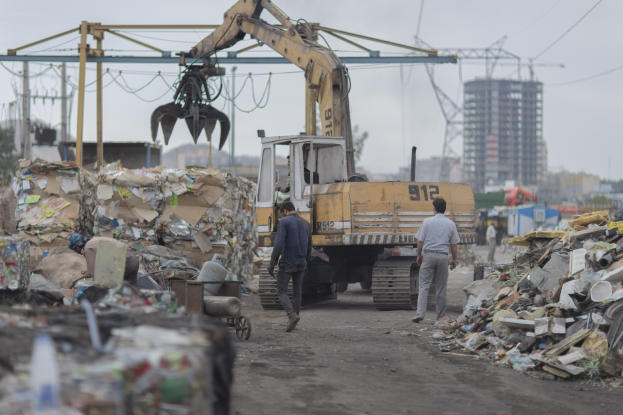 Eine Gruppe von Männern geht auf einer Schotterstraße neben einem Haufen Müll, mit einem Bagger im Hintergrund, Gebäuden, Bäumen, Polen und Drähten, die in der Gegend verstreut sind.