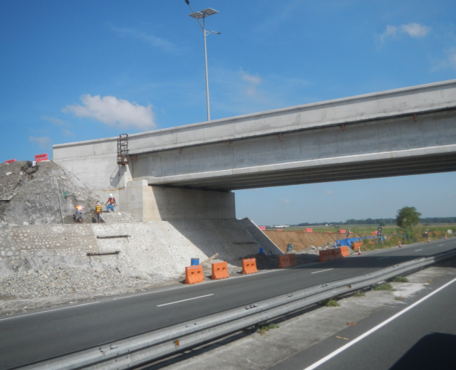 Autobahn mit einer Brücke darüber, ein darunter vorbeifahrendes Auto und Bauarbeiter am Straßenrand, umgeben von Verkehrskegeln, Steinen und einem Lichtmast, mit Bäumen und einem bewölkten Himmel im Hintergrund.
