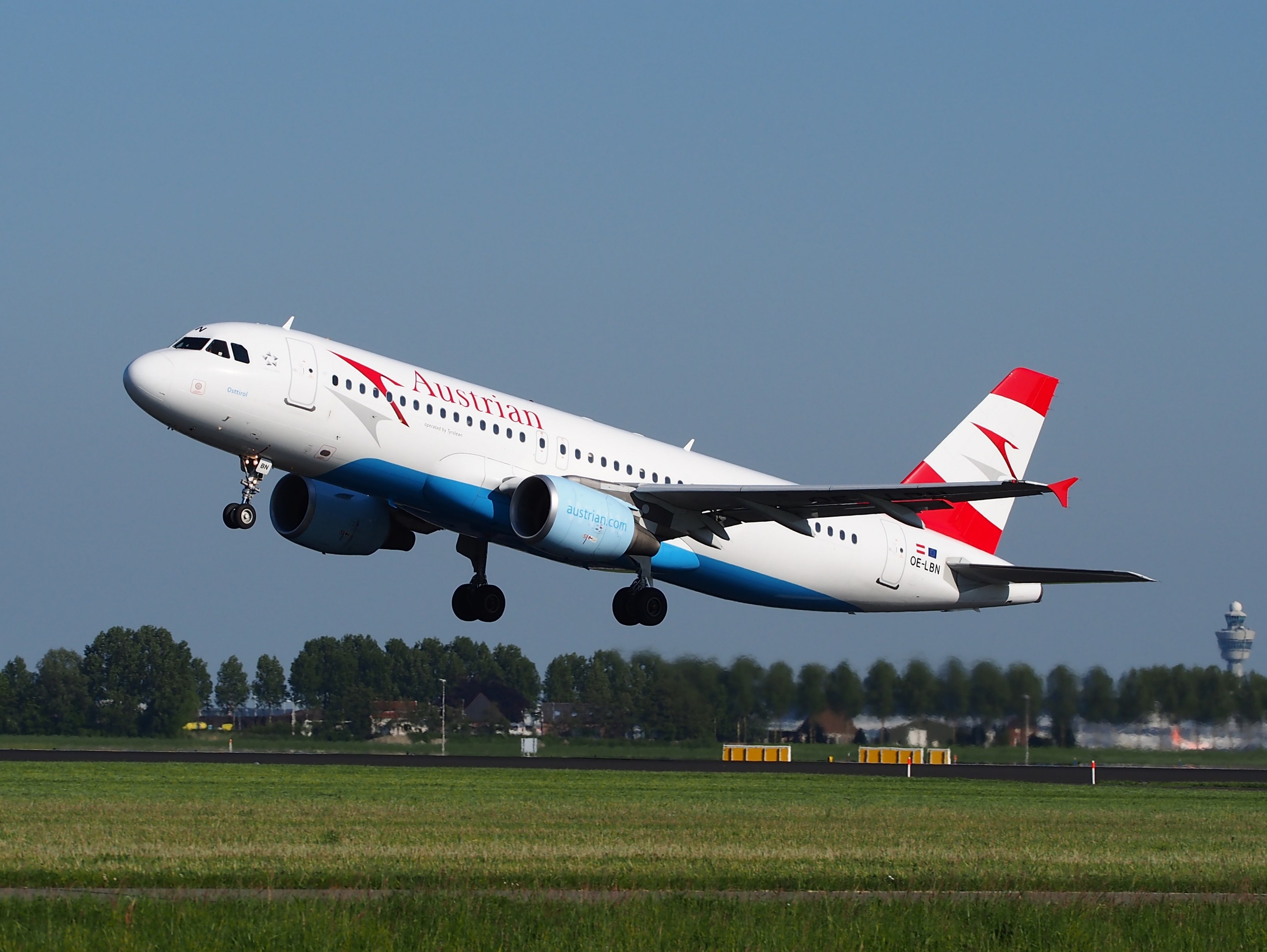 Airbus A320-200 der Austrian Airlines beim Start vom Frankfurter Flughafen, mit grünem Gras, Bäumen, Gebäuden und einer Kontrollturm im Hintergrund.