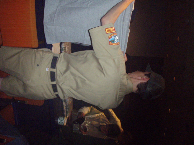 Ein Polizist hält ein T-Shirt in den Vordergrund, mit einem Jungen im Hintergrund, der ein Buch und einen Becher hält, neben einem Schreibtisch und Lampen.