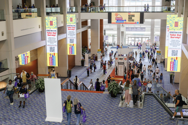 Eine große Gruppe von Menschen, die durch ein großes Gebäude mit Bannern, Topfpflanzen, Rolltreppen, Geländern, Säulen, bedruckten Tafeln, Glasdoors und anderen Gegenständen geht, einige mit Taschen, wahrscheinlich beim Nationalen Buchfestival.