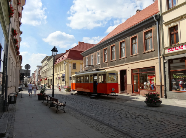 Eine rote und gelbe Straßenbahn fährt auf einem Kopfsteinpflasterweg durch die Altstadt von Tallinn, flankiert von Gebäuden mit Fenstern, Bänken und Laternen, mit Passanten auf den Gehwegen und einem bewölkten Himmel im Hintergrund.