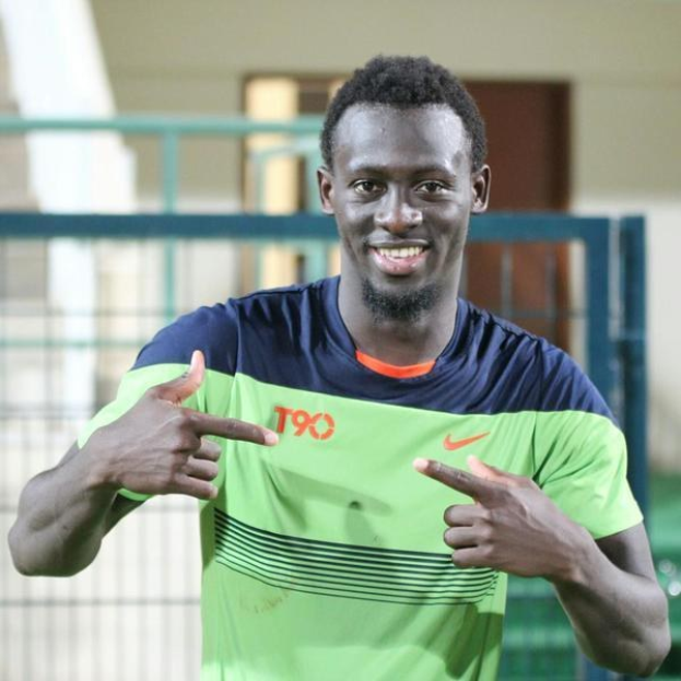 Ein Mann in einem grün-blauen Hemd, der in die Kamera lächelt und zeigt, vor einem unscharfen Hintergrund mit einem Geländer, einer Wand und einer Tür.