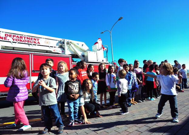 Kinder vor einem Feuerwehrauto mit der Aufschrift 'Yokosuka Fire Department' bei einer Weihnachtsparade, umgeben von Laternenmasten, Bäumen und einem klaren blauen Himmel.