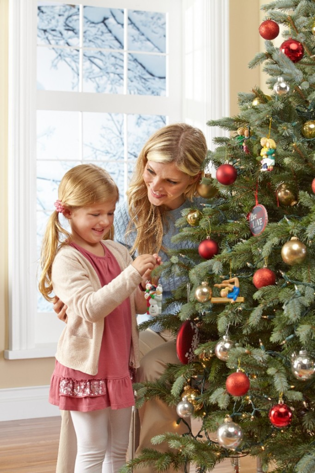 Ein geschmückter Weihnachtsbaum mit einer Frau und einem Kind davor, ein Fenster und eine Wand im Hintergrund und Äste vor dem Fenster.