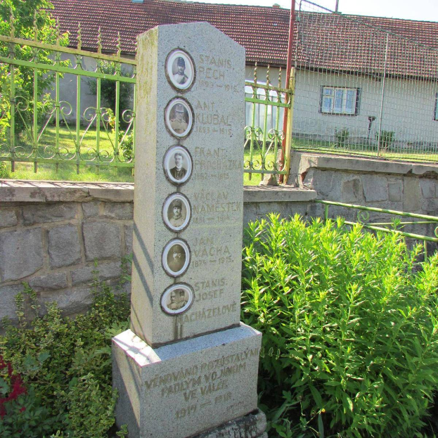 Ein Steindenkmal in einem Garten, umgeben von Pflanzen, einer Wand, einem Metallzaun, einem Haus, Pfählen, Gras und Bäumen unter einem bewölkten Himmel, zeigt Bilder und Text, die wahrscheinlich Holocaust-Opfern gewidmet sind.