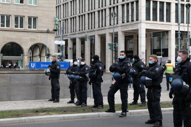 Eine Gruppe von Polizisten in schwarzen Uniformen und Masken steht vor einem Berliner Gebäude mit Glasfenstern und Säulen, einige halten Helme, mit Laternenmasten, Verkehrsampeln, Texttafeln, einer Statue und anderen Menschen im Hintergrund auf einer grünen Fläche.