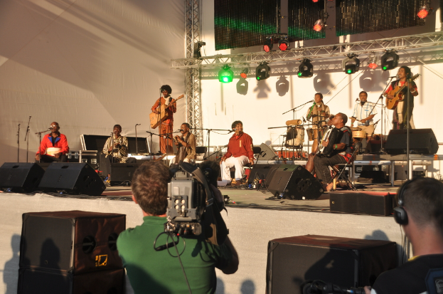 Ein musikalisches Konzert mit verschiedenen Instrumenten auf der Bühne, darunter Gitarren und Trommeln, unter bunten roten und grünen Scheinwerfern, mit Lautsprechern vorne und zwei Personen unten, eine filmt und die andere hört zu.