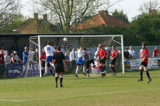 Fußballspieler sind in ein Fußballspiel auf einem Feld mit einem Tornetz involviert, während Zuschauer dahinter stehen, mit Bäumen und Häusern im Hintergrund sichtbar.