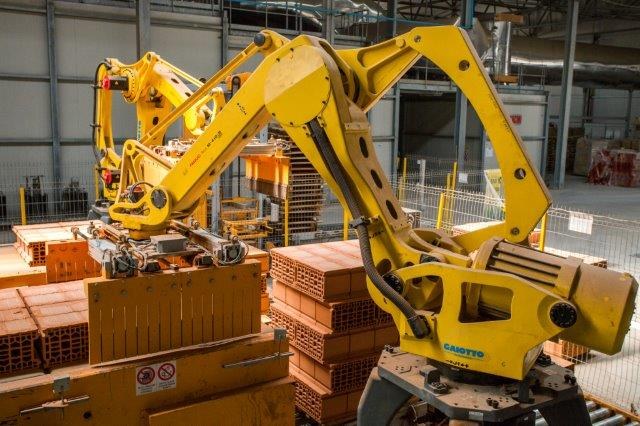 Großes Industriegebäude mit gestapelten Kisten, Kränen an der Decke, Zaun, Pfählen und einer fernen Wand in einer Fertigungseinrichtung.