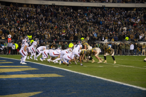 Ein Fußballspiel im Gange auf einem Stadionfeld mit Spielern in Sportbekleidung und Helmen auf dem Boden, während Zuschauer und Stadionmerkmale im Hintergrund zu sehen sind.