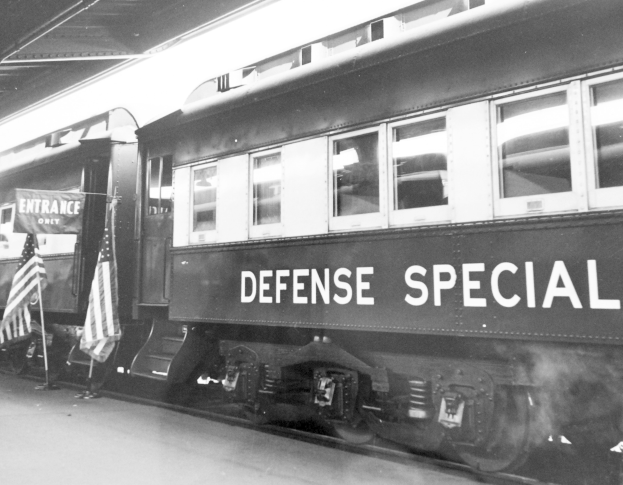 Schwarz-weiss-Foto eines Zuges mit der Aufschrift "Defense Special" auf den Eisenbahnschienen mit zwei Fahnen links und einer Decke über.