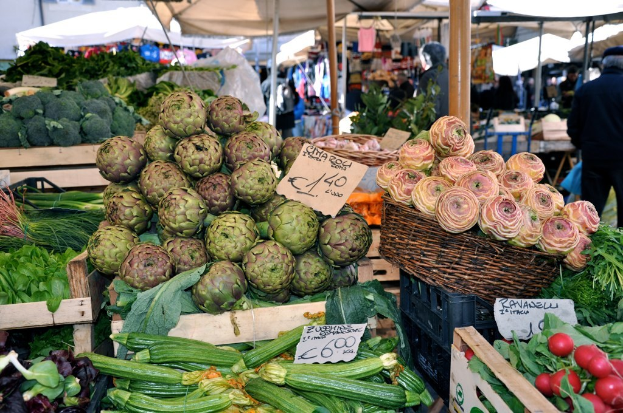 Körbe gefüllt mit Obst, Gemüse und kleinen Schildern mit Text, mit Zelten und ein paar Menschen im Hintergrund.