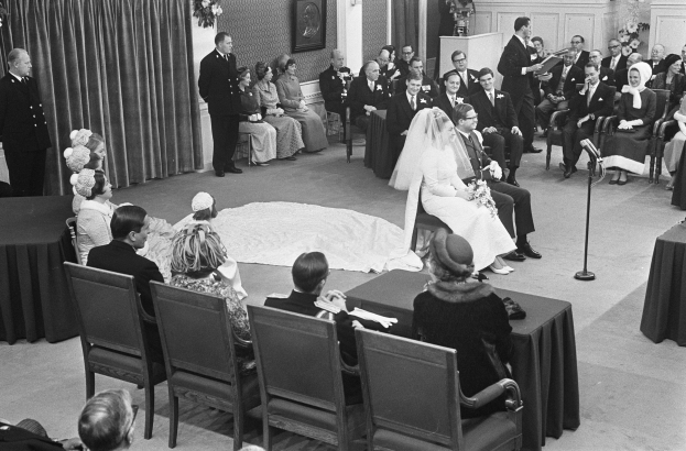 Schwarzes und weißes Foto einer Hochzeitszeremonie mit sitzenden und stehenden Menschen, einem Mann, der in ein Mikrofon spricht, und einer Frau, die einen Blumenstrauß hält, mit einem Podium, Blumengestecken, einem Vorhang, einem gerahmten Foto an der Wand und einer Tür im Hintergrund.