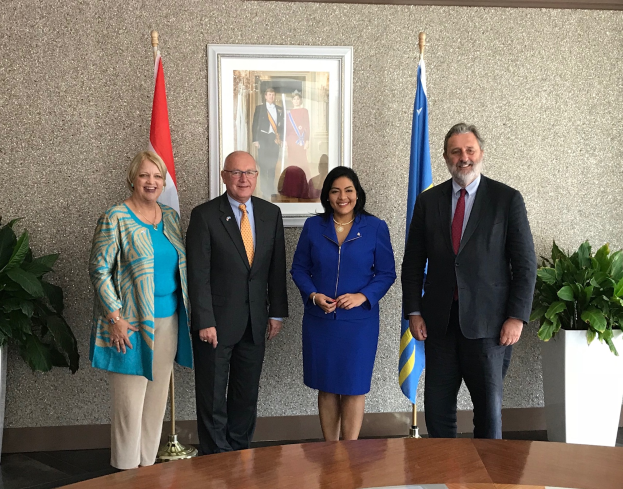 Eine Gruppe strahlender Menschen um einen Tisch mit Papieren herumstehend, umgeben von Fahnen, Topfpflanzen und einem Foto auf der Wand, feiernd die Unterzeichnung eines Memorandums zwischen den Vereinigten Staaten und Kanada.