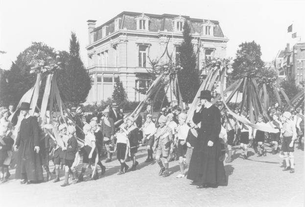 Ein Schwarz-Weiß- Bild einer Gruppe von Menschen, die mit Fahnen in der Hand die Straße entlanggehen, mit Hüten auf dem Kopf, vor einem Gebäude, Bäumen und einem sichtbaren Himmel, mit einem Maibaum im Hintergrund.