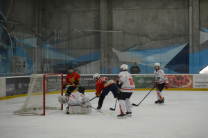 Gruppe von Menschen, die Eisstockschüßen auf einem Eisplatz spielen, mit Helmen und Eishockeyschläger, mit einem Tor auf der linken Seite, Bannern im Hintergrund und einer Wand.