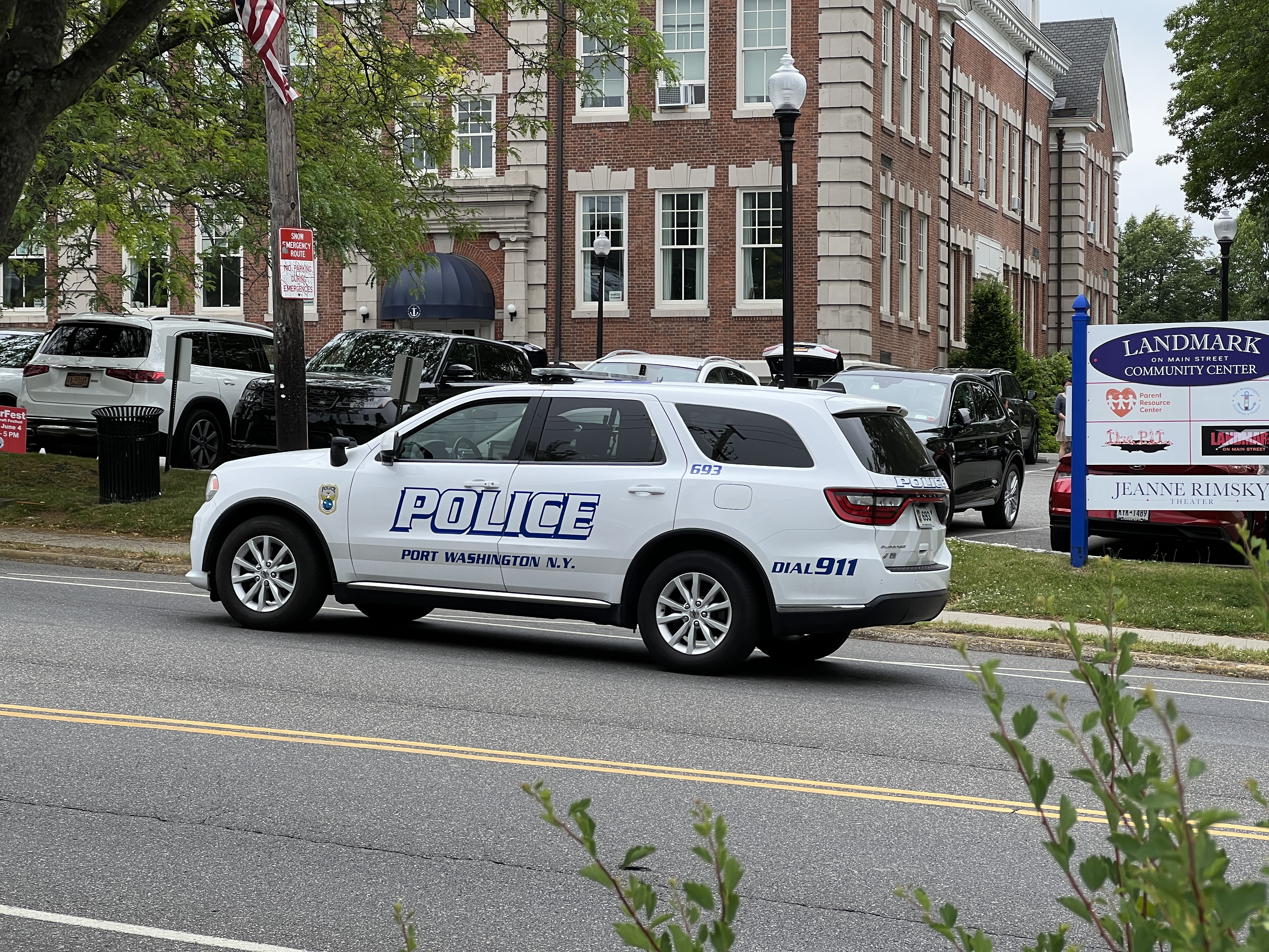 Ein Polizeiwagen auf der Seite der Straße vor einem Gebäude mit Fenstern, Laternenmasten, Schildern, einer Fahne, Bäumen und einem Müllcontainer im Vordergrund, unter einem bewölkten Himmel.