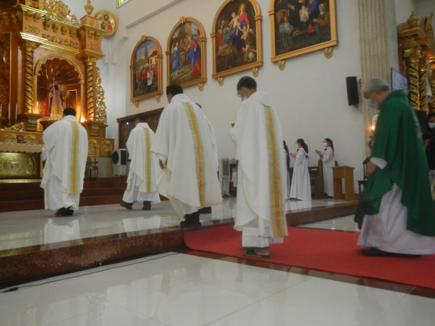 Eine Gruppe von Priestern, die auf einem roten Teppich in einer Kirche entlanggehen, mit Menschen, die vor ihnen auf dem Boden stehen und auf der rechten Seite, und Fotorahmen, die an der Wand im Hintergrund befestigt sind.