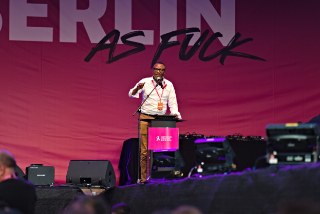 Ein Mann in einem weißen Hemd und braunen Hosen steht auf einer Bühne und hält ein Mikrofon, mit einem pinken Banner mit der Aufschrift 'Berlin as Fuck' dahinter und Lautsprechern auf der Bühne.