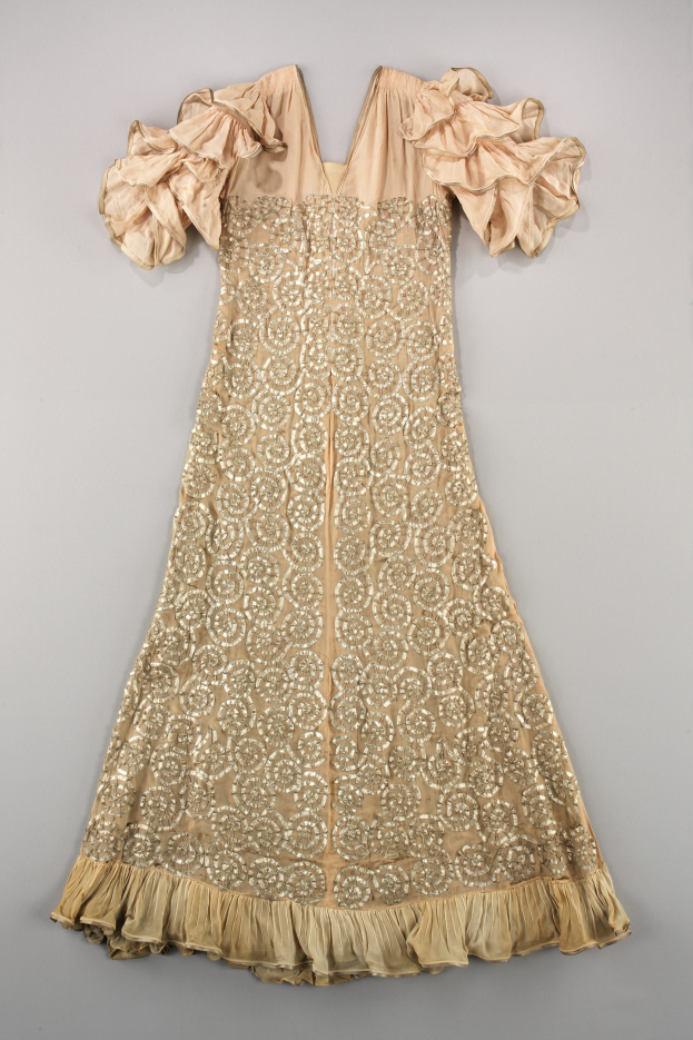 Ein cremefarbenes Kleid mit goldener Stickerei und Rüschen, das auf einer weißen Oberfläche detaillierte Designs zeigt.