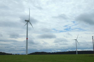 Drei hohe weiße Windräder in einem grünen Feld mit Bäumen und Wolken im Hintergrund.
