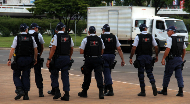 Eine Gruppe von Beamten in Uniform, einschließlich Mützen, Hemden, kugelsicheren Westen, Uhren, Hosen und Schuhen, geht auf einer Straße. Rechts daneben steht ein Lastwagen in der Nähe eines Zauns, mit Gebäuden, Bäumen, Pflanzen, Polen und Bannern im Hintergrund. Eine Brücke ist oben rechts zu sehen.