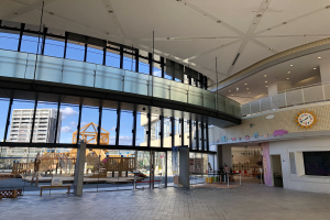 Innenansicht eines großen Gebäudes mit hohen Fenstern, Deckenleuchten, einer Wanduhr, einem Fernseher, einem Anzeigebildschirm, hölzernen Strukturen, einem hölzernen Zaun, entfernten Gebäuden, Bäumen und einem bewölkten Himmel, die den Eingang zu einem neuen Kinderspielplatz markieren.