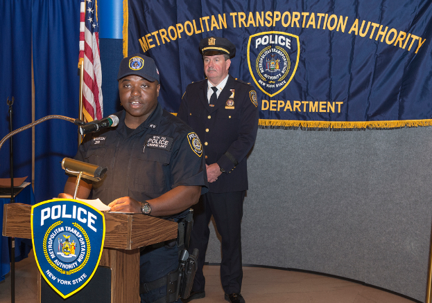 Zwei Polizisten stehen vor einem Rednerpult, einer hält Papiere und spricht in ein Mikrofon, mit einer Fahne, einem Tisch und einer Tafel im Hintergrund.