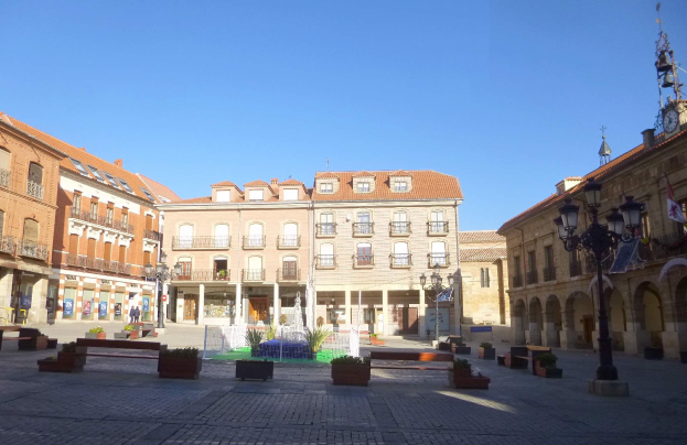Ein Rathausplatz in einem Stadtplatz mit einem zentralen Brunnen, umgeben von Bänken, Topfpflanzen, Straßenlaternen, einem Uhrenturm und Gebäuden mit Fenstern unter einem klaren blauen Himmel.