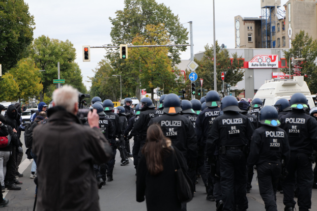 Eine Gruppe von Menschen geht über eine Straße mit Polizeibeamten, einige tragen Helme und haben Taschen oder halten Kameras, mit Fahrzeugen, Verkehrszeichen, Schildern, Bäumen, Gebäuden und einem klaren Himmel im Hintergrund.