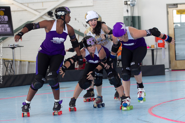 Vier Personen mit Helmen fahren in der Mitte, mit einer Wand, Tür, Ständer, Mikrofon, Schildern, Tisch und anderen Gegenständen im Hintergrund.