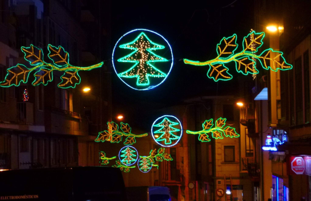 Eine festlich geschmückte Stadtstraße bei Nacht, dekoriert mit Weihnachtslichtern in Form von Blättern und anderen Schmuckelementen, mit Fahrzeugen, die auf beiden Seiten an Gebäuden vorbeifahren.