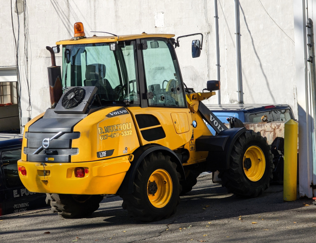 Ein gelber Radlader auf einer Straße vor einem Gebäude mit einer Wand im Hintergrund und anderen Fahrzeugen in der Nähe geparkt.