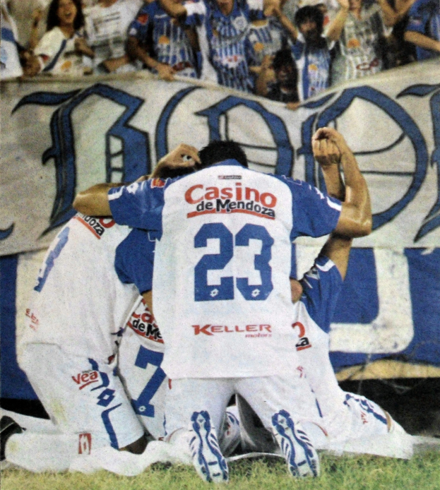 Eine Gruppe von Fußballspielern feiert ein Tor auf dem Boden vor einer jubelnden Menge, mit einem Banner mit Text im Hintergrund.