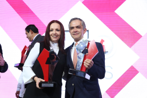 Zwei Menschen, die auf einer Bühne lächeln und Pokale halten, mit einem Bildschirm im Hintergrund.