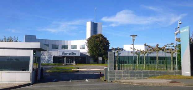 Ein großes Gebäude mit zahlreichen Fenstern und einem Tor davor, umgeben von Straßenmasten, Straßenlaternen, Gittern, Bäumen und Gras, mit einem bewölkten Himmel im Hintergrund, das als Hauptsitz von Iberostica in den Niederlanden dient.