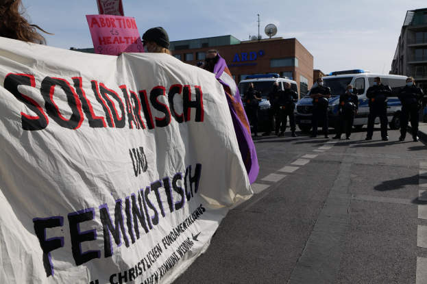 Eine Gruppe von Menschen marschiert auf einer Straße, hält ein Banner mit der Aufschrift 'Solidarität und Feminismus', mit geparkten Fahrzeugen und Gebäuden im Hintergrund unter einem klaren blauen Himmel.