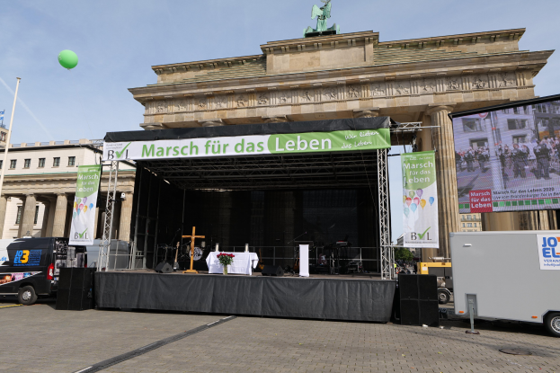 Bühne vor dem Brandenburger Tor mit einem Tisch, Bannern, Lautsprechern, Fahrzeugen, Gebäuden, einer Statue, einem Fahnenmast und bewölktem Himmel.