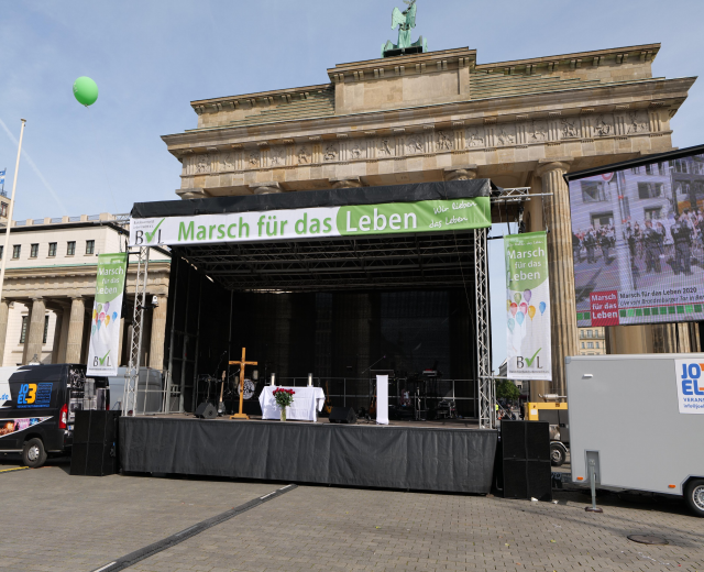 Bühne vor dem Brandenburger Tor mit einem Tisch, Bannern, Lautsprechern, Fahrzeugen, Gebäuden, einer Statue, einem Fahnenmast und bewölktem Himmel.