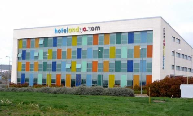 Großes modernes Hotelgebäude mit vielen Fenstern, umgeben von Grün, einem Zaun, Straßenlaternen, geparkten Fahrzeugen, fernen Hügeln und einem bewölkten Himmel; das Wort "hotel and go" ist auf dem Gebäude zu sehen.