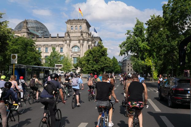 Eine Gruppe von Menschen, die auf Fahrrädern eine von Bäumen gesäumte Straße in Berlin, Deutschland, entlangfahren, mit Gebäuden und einer Bushaltestelle auf der rechten Seite, unter einem bewölkten Himmel und einer Flagge auf einem der Gebäude.